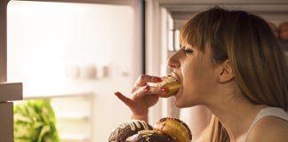 Close up image of a young woman with eating disorder, having a midnight snack - eating donuts, in front of the refrigerator.